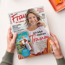 Aufgeschlagene Zeitschrift „Frau im Leben“ wird von zwei Händen über einem weißen Tisch gehalten; auf dem Cover eine lächelnde blonde Frau in weißer Bluse. Sichtbare Titel: „Ich lebe meinen Traum“, „10 Ideen für kleine Räume“, „Neue Mode – Die Trends im Herbst und Winter“, „Die 5-Euro-Küche“ und „Bayern für Entdecker“; oben rechts liegt ein orangefarbenes Objekt.