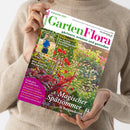 Person in beigefarbenem Strickpullover hält eine schräg nach rechts geneigte Gartenzeitschrift mit weißem Cover; darauf ein farbenfrohes Blumenbeet und ein kleines Foto einer Frau im Garten. Lesbare Texte: „GartenFlora“, „September 2026“, „gärtnern, erleben und genießen“, „Unkompliziert Mini-Kiwis für den Garten“ und „Magischer Spätsommer in Burgunderrot“.