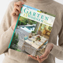 Eine weibliche Person im beigefarbenen Strickpullover hält schräg eine Zeitschrift mit türkisfarbenem Rand vor hellem Hintergrund. Das Cover zeigt eine begrünte Terrasse mit Pool, gedecktem Esstisch und Wohnbereich; lesbar sind „GARTEN & WOHNEN“, „Wohnträume“ und „Genießen wie im sonnigen Süden“ sowie Hinweise auf „Cloud Dancer“, „Provence“, „Côte d’Azur“ und „Lavendelfeldern“.