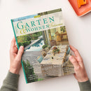 Hochformatige Zeitschrift mit türkisfarbenem Rand, von zwei Händen über einer weißen Fläche gehalten; auf dem Cover stehen „GARTEN“, „WOHNEN“ und „Träume“ sowie „Genießen wie im sonnigen Süden“. Die Collage zeigt einen Garten mit Pool, eine gedeckte Terrasse und einen Innenraum; oben rechts liegt ein orangefarbenes Objekt.