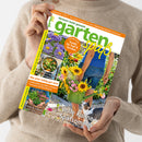 Weibliche Person im beigen Strickpullover hält ein schräg abgebildetes Gartenmagazin mit dem Titel „gartenspaß“ und der Überschrift „Gärtnern leicht gemacht“. Das Cover zeigt eine Person mit grüner Gießkanne, Sonnenblumen und Blumen, eine Katze sowie Teasertexte wie „Blühende Astern in allen Farben“, „Mal anders kochen mit Asia-Blattgemüse“ und „Schick & schnell Pflanzen-Deko im Korb“.