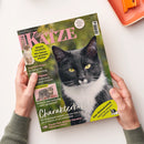 Schräg auf einer weißen Fläche gehaltenes Magazin „Geliebte KATZE“ mit großem Schwarz-Weiß-Foto einer Katze, gelben Augen und weißer Brust sowie zwei kleineren Katzenabbildungen. Lesbare Texte: „Europas beliebtestes Katzenmagazin“, „Aktuelle Forschung: Wie Katzen uns gesünder machen“, „Charakterkopf“ und „Zum Weltkatzentag“; zwei Hände halten das Magazin, oben rechts liegt ein orangefarbenes Objekt.