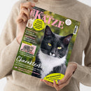 Eine Person im beigefarbenen Strickpullover hält ein schräg ausgerichtetes Magazin mit dem Titel „Geliebte Katze“, Ausgabe August 2026, vor weißem Hintergrund. Das Cover zeigt eine schwarz-weiße Katze, ein Kätzchen und ein graues Tabby-Katzenfoto sowie die Texte „Aktuelle Forschung: Wie Katzen uns gesünder machen“, „Experten-Tipps“, „Charakterkopf“ und „Zum Weltkatzentag“.