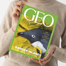 Person im hellbeigen Strickpullover hält ein schräg ausgerichtetes grünes GEO-Magazin mit dem Titel „GEO“ und dem Text „DIE WELT MIT ANDEREN AUGEN SEHEN“. Das Cover zeigt einen Pinguin in gelber Kleidung sowie die Aufschriften „Expedition zu den DANGER ISLANDS“, „Deutschlands weißer Fleck am Ende der Welt“, „KÄFER“, „SOKRATES“ und „MEXIKO“.