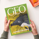 Grünes GEO-Magazin „AUGUST 2020“ in schräger Draufsicht, von zwei Händen gehalten. Das Cover zeigt einen schwarz-weißen Pinguin in gelber Kleidung und enthält die Texte „DIE WELT MIT ANDEREN AUGEN SEHEN“, „KÄFER“, „SOKRATES“, „MEXIKO“ sowie „Expedition zu den DANGER ISLANDS“ und „Deutschlands weißer Fleck am Ende der Welt“; oben rechts liegt teilweise ein orangefarbenes Etui.