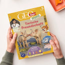 Gelbes GEOlino-mini-Magazin mit dem Titel „Tierische Freundschaft“, schräg auf weißem Untergrund liegend und von zwei Händen gehalten; auf dem Cover sind zwei Schweine, ein gezeichnetes Tiermaskottchen und ein Bergfoto abgebildet. Sichtbare Texte: „Die Welt für junge Entdeckerinnen und Entdecker“, „ab 5 Jahren“, „Kinder Manuela lebt im Regenwald“, „Basteln: Lass die bunten Puppen tanzen“ und „Wissen: Hoch hinaus – so entstehen Berge“.