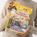 Gelbes GEOlino-mini-Magazin „Tierische Freundschaft“ in den Händen einer Person mit beigem Strickpullover. Covertexte: „Die Welt für junge Entdeckerinnen und Entdecker“, „ab 5 Jahren“, „Kinder: Manuela lebt im Regenwald“, „Basteln: Lass die bunten Puppen tanzen“ und „Wissen: Hoch hinaus: So entstehen Berge“; abgebildet sind zwei Ferkel, ein Bergfoto und ein gezeichneter Leopard.