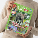 Hände einer nicht näher erkennbaren Person im beigefarbenen Strickpullover halten schräg eine grüne GEOlino-Zeitschrift. Das Cover zeigt drei Elefanten im Wasser, einen Mann bei „Checker TOBI“, ein Satellitenmotiv und Texte wie „Die Welt für junge Entdeckerinnen und Entdecker“, „TIERISCHE KLIMASCHÜTZER“ sowie „Was Waldelefanten, Biber und Wale für die Umwelt tun“.