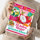 Hochformatiges, glänzendes Magazin „Good Health“ in den Händen einer Person ohne erkennbares Gesicht, vor einem beigefarbenen Strickpullover. Das pink-weiße Cover zeigt eine aufgeschnittene Drachenfrucht, Sanddornbeeren, eine springende Frau und Brokkoli; sichtbare Texte: „12 Jahre länger gesund leben“, „Endlich schmerzfrei!“, „Japan-Workout“ und „Kraftpaket Brokkoli“.