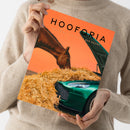 Person in beigefarbenem Strickpullover hält eine orangefarbene Zeitschrift mit dem sichtbaren Titel „HOOFORIA“ und dem Text „… DIE KUNST DER PAUSE“. Das Cover zeigt ein braunes Pferd, einen grünen Oldtimer und Stroh; die Zeitschrift ist schräg aus der Aufsicht fotografiert.