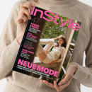 Eine Person in beigefarbenem Strickpullover hält ein schräg ausgerichtetes Magazin mit schwarzem Cover und pink-weißen Überschriften. Lesbar sind „in style“, „TREND-SHOES“, „SKIN-TONE HAIR“ und „NEUE MODE“; das Cover zeigt eine Frau in heller Kleidung vor einem Fenster sowie Bäume im Hintergrund.