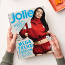 Auf weißer Fläche hält eine Person mit beiden Händen ein schräg liegendes Magazin der Marke „jolie“ mit dem Text „jolie.de“ und „MEGA TREND EDITION“. Das Cover zeigt eine weibliche Person mit langem schwarzem Haar in roter Jacke und roter Hose; sichtbar sind außerdem „BEST OF BEAUTY“, „10 geniale Tipps“ sowie „Leder, Seventies, Denim, Strick, Sporty Layering“.