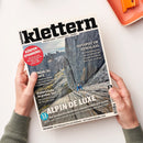 Schräg von oben fotografierte Zeitschrift „Klettern“ auf weißer Fläche, gehalten von zwei Händen mit grauen Strickärmeln; oben rechts liegt ein orangefarbenes rechteckiges Objekt. Das Cover zeigt eine felsige Berglandschaft mit einer kletternden Person und trägt die Texte „ABENTEUER | LIFESTYLE | SPORT“, „HOTSPOT IM HIMALAJA“, „KÖRPER-SPANNUNG“, „Kletterschuhe im großen Test“ und „ALPIN DELUXE“.