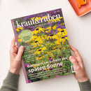 Schräg gehaltenes Magazin „kraut&rüben“ mit violettem Rand und Titelbild gelber Blüten mit dunklen Blütenmitten vor violett-grünem Hintergrund. Lesbare Texte: „SCHÖNER. WILDER. BIOGARTEN.“, „COOL-FLOWER-METHODE: Jetzt säen, im Frühling strahlen“, „Gärtnern im Schein der späten Sonne“ und „Die Tomaten sind reif!“; zwei Hände halten das Magazin über einer hellen Fläche, oben rechts liegt ein orangefarbenes Notizbuch mit Klammer.