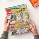 Auf weißer Fläche halten zwei Hände eine schräg liegende, glänzende Wohnzeitschrift mit türkisfarbenem, gelbem, rotem und violettem Cover. Lesbar sind „Lisa Wohnen & Dekorieren“, „Kreativ dem Sommer verlängern“, „EXTRA 60 Ideen für ein leichtes Wohngefühl“, „Landhaus-Liebe“ und „Natürlich schön: Dekorieren mit Blumen, Gräsern & Co.“; abgebildet sind ein helles Esszimmer, Blumen, eine Frau und Gebäck.