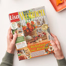 Auf weißer Fläche halten zwei Hände eine schräg liegende Zeitschrift „Lisa Wohnen & Dekorieren“ mit orangefarbenem Herbstmotiv, Blumen, Weidenkorb, Kürbissen und Dekoration. Lesbare Texte: „58 kreative Herbst-Ideen“, „Wohlfühl-Mix Orange und Naturtöne“, „Dekolust im Herbst“ sowie „Die besten Kürbis-Rezepte“; oben rechts liegt ein orangefarbenes Tablett.