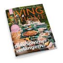 Schräg fotografiertes Hochglanzcover der Zeitschrift „LIVING AT HOME“ mit dem Schriftzug „Mein Zuhause voller Ideen“. Eine gedeckte Tafel mit türkisfarbener Tischdecke, Geschirr, Gläsern, Tassen, Kuchen, Obst, Blumen und Korbstuhl vor grünem Gartenhintergrund; sichtbare Texte: „GUTE LAUNE IN DER KÜCHE“, „Süß bis herzhaft“, „CITYTRIP PORTO“, „Spätsommer-Glück“ und „Der Sommer verändert“.