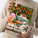 Weibliche Person im beigefarbenen Strickpullover hält schräg eine Zeitschrift vor hellem Hintergrund. Das Cover zeigt ein gedecktes Gartentisch-Arrangement mit Speisen, Geschirr, Blumen und Grün sowie die Texte „LIVING AT HOME“, „Mein Zuhause voller Ideen“, „GUTE LAUNE IN DER KÜCHE“, „CITYTRIP PORTO“ und „Den Sommer verlängern“.