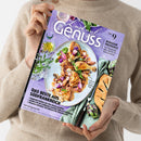 Eine Person im beigen Strickpullover hält ein schräg ausgerichtetes Magazin mit violettem Cover. Sichtbar sind der Titel „Lust auf Genuss“, „Das Beste aus Südfrankreich“, „Bonjour Provence“ sowie ein Teller mit gebratenem Geflügel, Gemüse und Kräutern; daneben liegen Brot, Artischocken und Besteck auf grünem Stoff.