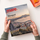 Zwei Hände in grauen Strickärmeln halten ein schräg liegendes Magazin über einer weißen Oberfläche. Auf dem Cover stehen „mare“, „Die Zeitschrift der Meere“, „Nepal – Königin des Südens“ sowie „Deep Talk: Wie Wale telefonieren“, „Mississippi-Delta: Der Blues der Cajuns“ und „‚Cap Arcona‘: Die vergessene Katastrophe in der Lübecker Bucht“; abgebildet sind eine Küstenstadt, eine Bucht und Berge bei Sonnenuntergang.