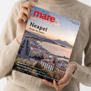 Eine weibliche Person im cremefarbenen Strickpullover hält eine schräg abgebildete Zeitschrift mit dem roten Titel „mare“ und dem Untertitel „Die Zeitschrift der Meere“. Das Cover zeigt eine Küstenstadt am Meer bei Sonnenuntergang, mit den Texten „Neapel“, „Königin des Südens“ sowie weiteren Themenhinweisen zu Walen, dem Mississippi-Delta, „Cap Arcona“ und der Lübecker Bucht.