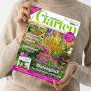 Person mit beige gestricktem Pullover hält ein schräg abgebildetes Gartenmagazin mit dem Titel „Mein schöner Garten“. Das Cover zeigt einen blühenden Garten mit violetten und gelben Blumen sowie Texten wie „Zucchini & Kürbis“, „Klimaschutz + Artenvielfalt“, „Kein Garten ohne Hecke“ und „Blütenglück mit pflegeleichten Fetthennen“.