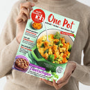 Weibliche Person im beigefarbenen Strickpullover hält ein schräg fotografiertes Kochmagazin. Das mintgrüne Cover von „meine Familie & ich“ trägt den Titel „One Pot“ und zeigt einen grünen Topf mit Gemüsegericht, einen Kuchen sowie Gewürze; sichtbare Texte sind „Schnell • lecker • aus einem Topf“, „Neue Rezepte!“, „Sommerküche“, „EINMACHZEIT“ und „Zwetschgen“.