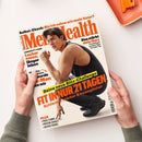 Aufgeschlagenes Magazin „Men’s Health“ mit weißem Cover, orange-schwarzer Typografie und gelbem Balken, gehalten von zwei Händen. Abgebildet ist eine männliche Person mit braunem Haar und schwarzem ärmellosem Oberteil in gebeugter Laufhaltung; sichtbare Texte: „Selbst-Check: Bin ich schon wie mein Vater?“, „Das stärkt dein Herz“, „Lecker essen, länger leben“, „Deine neue Blitz-Challenge: FIT IN NUR 21 TAGEN“ und „PLUS: Padel-Tennis, Protein-Shakes, Jacken-Trends“.