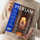 Diagonal gehaltenes Magazincover „MERIAN – The Art of Travel“ mit braunem Architekturmotiv: steinerner Torbogen, dahinter eine Kuppel im orangefarbenen Abendlicht. Auf blauem Rand stehen „Albanien“, „Finnland“, „Starke Städte“, „Lüneburger Heide“ und „Istrien“; unten „OH EUROPA!“. Eine Person im beigefarbenen Strickpullover hält das Cover mit beiden Händen, das Gesicht ist nicht sichtbar.