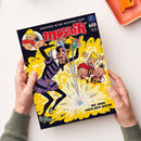 Mosaik-Magazin mit dunkelblauem, gelbem und rotem Cover, gehalten von zwei Händen einer nicht erkennbaren Person. Sichtbare Texte: „ABENTEUER IN DER GOLDENEN STADT“, „mosaik“ und „DIE JAGD NACH DEM GOLDE“; mehrere männlich dargestellte Comicfiguren vor gelbem Explosionshintergrund, daneben oben rechts ein orangefarbenes Tablett.