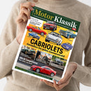 Hände einer weiblichen Person im beigefarbenen Strickpullover halten schräg eine Zeitschrift mit grün-gelbem Cover. Sichtbare Texte: „Motor Klassik“, „CABRIOLETS – Genießen Sie jeden Sonnenstrahl!“ sowie Fahrzeugbezeichnungen wie „Triumph Stag“, „Fiat Dino Spider“, „Porsche 911“, „Mercedes SLK“ und „Alfa 8C Competizione“; abgebildet sind mehrere klassische rote, blaue, weiße und grüne Automobile.