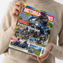 Hände einer Person im beigefarbenen Strickpullover halten eine schräg ausgerichtete Zeitschrift mit blau-rotem Cover. Sichtbare Texte: „MOTORRAD“, „Retro-Hornet“, „Fahrbericht: Honda CB 1000 F“, „KTM 790 Duke“, „Norton Atlas“ und „SOLO S 2000 GL“; abgebildet sind mehrere Motorräder mit fahrenden Personen sowie Landschaftsaufnahmen.