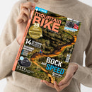 Weibliche Person im cremefarbenen Strickpullover hält eine schräg fotografierte Zeitschrift mit orange-blauem Cover vor hellem Hintergrund. Sichtbare Texte: „mountain BIKE“, „E-MTB“, „28 Seiten Reise“, „Kupplungsträger – Dreier-Träger im Test“, „14 XC-Reifen im Labor- & Praxistest“, „Bock auf Speed“ und „9 WM-Bikes im Test“; das Titelbild zeigt einen Mountainbike-Trail.