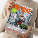 Eine Person ohne sichtbares Gesicht hält ein diagonal ausgerichtetes, glänzendes Fahrradmagazin vor einem beigefarbenen Strickpullover. Das Cover zeigt eine männliche Person mit Helm auf einem roten E-Bike vor modernen Gebäuden und trägt die Texte „E-BIKE“, „TOURENRAD-TEST UNTER 1800 EURO“, „BRANDNEU Specialized Vado 3X“ sowie „FAHRRAD-RUCKSÄCKE“ und „PANORAMA-TOUR IN DER SCHWEIZ“.