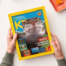 Gelb gerahmtes „NATIONAL GEOGRAPHIC“-Magazin mit frontal abgebildetem braun-weiß geflecktem Gecko, gehalten von zwei Händen einer nicht näher bestimmbaren Person. Lesbare Texte: „COOLE FAKTEN“, „verrückt, aber wahr!“, „GECKOS: Kopfüber auf Insektenjagd!“, „SCHIFFSWRACKS: Schätze unter Wasser!“, „SOMMER“ und „SCHOKOLADE“ sowie „2 tolle POSTER“.