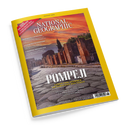 Schräg fotografiertes Magazin „NATIONAL GEOGRAPHIC“ mit gelbem Rand und dem Titel „POMPEJI“. Das Cover zeigt Ruinen, eine gepflasterte Straße und einen orangefarbenen Himmel; lesbare Texte: „AUGUST 2026“, „LEBEN UND STERBEN IM SCHATTEN DES VESUV“, „MAROKKOS AMAZONEN“, „MYSTERIÖSE BÄUMKREISE“ und „FLIEGENDE FRÜHMELDER“.