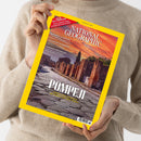 Eine Person ohne sichtbares Gesicht hält ein schräg fotografiertes Magazin mit gelbem Rand über einem beigefarbenen Strickpullover. Das Cover trägt den Titel „NATIONAL GEOGRAPHIC“, die Überschrift „POMPEJI – LEBEN UND STERBEN IM SCHATTEN DES VESUV“ sowie „AUGUST 2026“ und zeigt Ruinen bei Sonnenuntergang; weitere Texte nennen „MAROKKOS AMAZONE“, „MYSTERIÖSE BAUMKRISE“ und „FLIEGENDE FRÜHMELDER“.