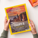 Gelb gerahmtes, schräg gehaltenes Magazin „NATIONAL GEOGRAPHIC“, Ausgabe „AUGUST 2026“, mit dem Titel „POMPEJI“ und dem Coverfoto antiker Ruinen bei orange-rotem Himmel. Sichtbare Texte: „MAROKKOS AMAZONEN“, „MYSTERIÖSE BAUMKREISE“ und „FLIEGENDE FRÜHMELDER“; zwei Hände einer nicht näher bestimmbaren Person halten das Magazin über einer weißen Fläche, oben rechts liegt ein angeschnittenes orangefarbenes Objekt.
