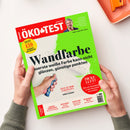 Schräg von oben fotografiertes ÖKO-TEST-Magazin mit rotem und grünem Cover, gehalten von zwei Händen mit grauen Ärmeln; oben rechts sind „ÖKO-TEST“ und „oekotest.de“ sowie ein Porträt und „INTERVIEW“ sichtbar. Haupttitel „Wandfarbe“, dazu Texte über weiße Farbe, Schadstoffe in der Raumluft und Junior-Zahnpasta; am Rand stehen Themen zu Erdbeer-Fruchtchips, Apfelessig, Fußpflege und Reinigungsmilch.