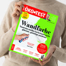 ÖKO-TEST-Magazin mit leuchtend grünem Rand und rotem Kopfbereich, schräg von oben fotografiert und von einer nicht näher erkennbaren Person im beigefarbenen Strickpullover gehalten. Das Cover zeigt weiße Wandfarbe und eine Hand mit blauem Farbroller sowie die Texte „Wandfarbe“, „Teuerste weiße Farbe kann nicht glänzen, günstige punkten“, „Dicke Luft?“ und „Test Junior-Zahnpasta“.