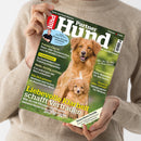 Weibliche Person im cremefarbenen Strickpullover hält eine schräg fotografierte Zeitschrift mit grünem Titelbild „Partner Hund“; darauf ein großer brauner Hund und ein Welpe vor Naturhintergrund. Sichtbare Texte: „Die Nr. 1 der Hunde-Magazine“, „Kleine Hunde erziehen“, „Fakten-Check“, „Schleppleine“, „Müde, durstig, schlapp“ und „Liebevolle Klarheit schafft Vertrauen“.