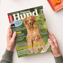 Schräg fotografierte Zeitschrift „Hundepartner“ auf weißer Fläche, gehalten von zwei Händen einer nicht näher bestimmbaren Person. Das grüne Cover zeigt einen braunen Hund und einen Welpen sowie Texte wie „Kleine Hunde erziehen“, „Osteopathie“, „Spielgruppen für Welpen“ und „Liebevolle Klarheit schafft Vertrauen“; oben rechts liegt ein orangefarbener Gegenstand.