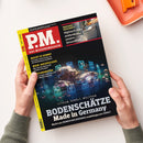 Schräg von oben fotografierte Zeitschrift „PM. Das Wissensmagazin“, die von zwei Händen gehalten wird. Schwarzes Cover mit rotem PM-Logo, gelbem Rand und den Texten „Grosses Geschäft: Hightech-Klos analysieren unsere Gesundheit“, „Macht KI dumm?“, „Wein-Archäologie“ sowie „Bodenschätze – Made in Germany“; darunter eine Abbildung von Gestein und chemischen Elementen. Im Hintergrund liegen ein oranges Notizbuch und eine orange Klammer.