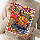 Weibliche Person im beigefarbenen Strickpullover hält eine schräg fotografierte Zeitschrift mit violettem Cover in beiden Händen. Lesbar sind „So ist das!“, „Für Ihre italienischen Momente“, „41 Saison-Hits auf italienische Art“, „Kürbis: 13-mal köstlich“, „Ofengerichte“ und „Aufläufe, Pasta & Co. Herbstgenuss“; abgebildet sind ein Nudelauflauf mit Hackbällchen und eine Frau mit roter Kochschürze.