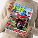 Weibliche Person im beigefarbenen Strickpullover hält ein schräg aufgenommenes Magazin „TOURENFAHRER“ mit blau-gelbem Titel, rotem Motorrad und weiteren Motorradbildern. Lesbare Texte: „REISERETTUNG: ERSTE HILFE BEI NOTFÄLLEN“, „TOUREN-TEST BMW F 450 GS Trophy“, „FAHRBERICHT SUZUKI SV-7GX“, „GROSSE GEFÜHLE ROYAL ENFIELD Classic 650“ und „MAXIMALBOXER“.