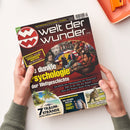 Zwei Hände halten ein schräg fotografiertes Magazin „Welt der Wunder“ mit schwarzem, rotem und gelbem Cover. Sichtbare Texte: „Entdecken–Staunen–Wissen“, „Enthüllt: aus den KRANKEN-AKTEN der Mächtigen“, „Die dunkle Psychologie der Weltgeschichte“, „7 TRAUM-STRÄNDE“, „Wie erkennt man ein DEEP-FAKE?“ und „Mit Sack und Pack“. Im Hintergrund liegen ein orangefarbenes Tablett und ein orangefarbener Gegenstand.