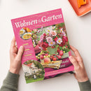 Rosa Zeitschrift „Wohnen & Garten“ mit dem Untertitel „Die schönsten Ideen für drinnen und draußen“, schräg von oben auf weißem Untergrund fotografiert und von zwei Händen mit grauen Ärmeln gehalten. Das Cover zeigt einen Blumenstrauß in Rosé, Obst und eine Suppenschüssel; lesbare Texte sind „Blüten-Finale in Rosé“, „Garten & Pflanzen“ und „Ländliches Spätsommerglück in Schweden“.