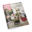 Schräg fotografiertes Cover der Wohnzeitschrift „WOHN IDEE“ vor schwarzem Hintergrund mit hellem Wohnzimmer, grünen Sesseln, weißem Sofa, Pflanzen, Couchtisch, Stehlampe und Wandbild. Sichtbare Texte: „Garten-Checkliste – Was Beete und Pflanzen jetzt brauchen“, „Bitte zu Tisch“, „Kühle Brise“, „Die pure LEICHTIGKEIT“ sowie „Pflanzen, Ordnung, Licht: Mit Ferien-Feeling und Feng-Shui zu mehr Balance und Harmonie zu Hause“.