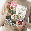 Eine Person in beigefarbenem Strickpullover hält schräg eine Zeitschrift mit rosa-weiß-grünem Cover und dem Titel „WOHNIDEE“. Abgebildet sind ein helles Wohnzimmer mit grünem Sofa, Pflanzen, Tisch und Sesseln; lesbare Texte: „Garten-Checkliste“, „Die pure LEICHTIGKEIT“, „BITTE ZU TISCH“, „KÜHLE BRISE“ und „Sommer-Gewinnspiel Heiße Preise“.