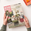 Schräg fotografierte Wohnzeitschrift „WOHN IDEE“, von zwei Händen einer nicht näher bestimmbaren Person gehalten; die Person trägt einen graugrünen Strickpullover. Das Cover zeigt ein helles Wohnzimmer mit grüner Sitzmöbelgarnitur, Pflanzen, Couchtisch und Leuchte sowie die Texte „Garten-Checkliste“, „Was Beete und Pflanzen jetzt brauchen“, „Die pure Leichtigkeit“ und „Der große Sofa-Guide“; oben rechts liegt ein orangefarbenes Schreibbrett.